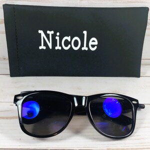 Pu&ograve; includere: Occhiali da sole neri con lenti colorate blu e una custodia nera con il nome "Nicole" stampato in bianco.