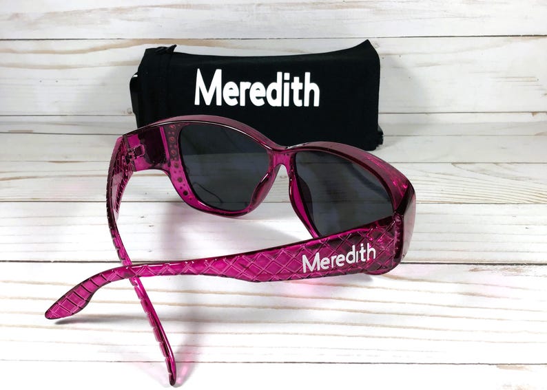 May include: Pink sunglasses with dark lenses and the name "Meredith" printed on the arm. A black case with the name "Meredith" in white is behind the sunglasses. The sunglasses have a textured pattern on the frames.