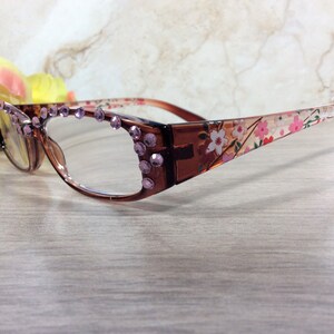 May include: Brown-framed spectacles with clear lenses. The frames are decorated with purple rhinestones and a floral pattern on the arms. The spectacles are displayed on a light-coloured wooden surface.