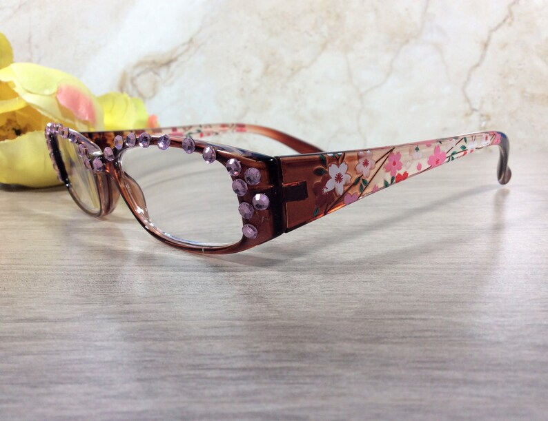 May include: Brown-framed spectacles with clear lenses. The frames are decorated with purple rhinestones and a floral pattern on the arms. The spectacles are displayed on a light-coloured wooden surface.