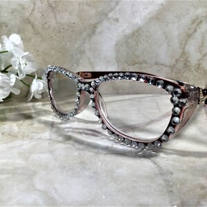 May include: Cat-eye style eyeglasses with a rose-gold frame, embellished with sparkling rhinestones along the top and outer edges. The glasses are set against a marble background, with a cluster of white flowers in the upper left corner.