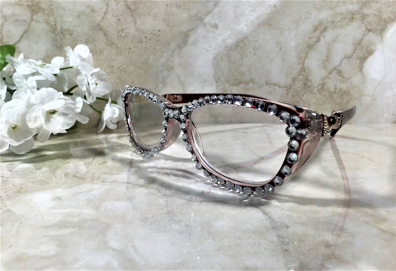 May include: Cat-eye style eyeglasses with a rose-gold frame, embellished with sparkling rhinestones along the top and outer edges. The glasses are set against a marble background, with a cluster of white flowers in the upper left corner.