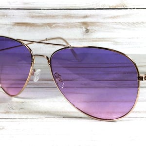 May include: Aviator sunglasses with a gold-coloured frame and gradient purple lenses. The lenses transition from a lighter pink at the bottom to a deep purple at the top. The sunglasses are set against a white wooden background.