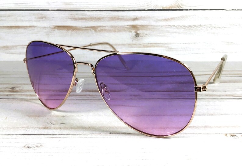 May include: Aviator sunglasses with a gold-coloured frame and gradient purple lenses. The lenses transition from a lighter pink at the bottom to a deep purple at the top. The sunglasses are set against a white wooden background.