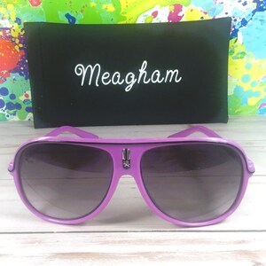 Puede incluir: Gafas de sol aviador moradas con lentes oscuras y un estuche negro. El estuche tiene el nombre "Meagham" en escritura blanca. Las gafas tienen un emblema plateado en el puente, sobre un fondo colorido.