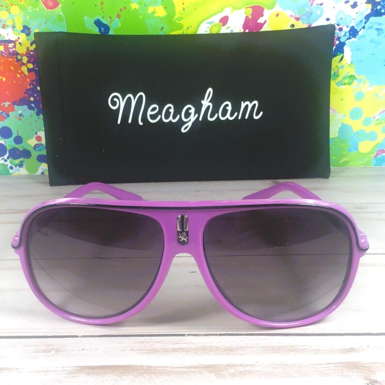 Puede incluir: Gafas de sol aviador moradas con lentes oscuras y un estuche negro. El estuche tiene el nombre "Meagham" en escritura blanca. Las gafas tienen un emblema plateado en el puente, sobre un fondo colorido.