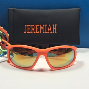 Puede incluir: Gafas de sol naranjas con lentes espejados y una funda negra con el nombre "JEREMIAH" bordado.