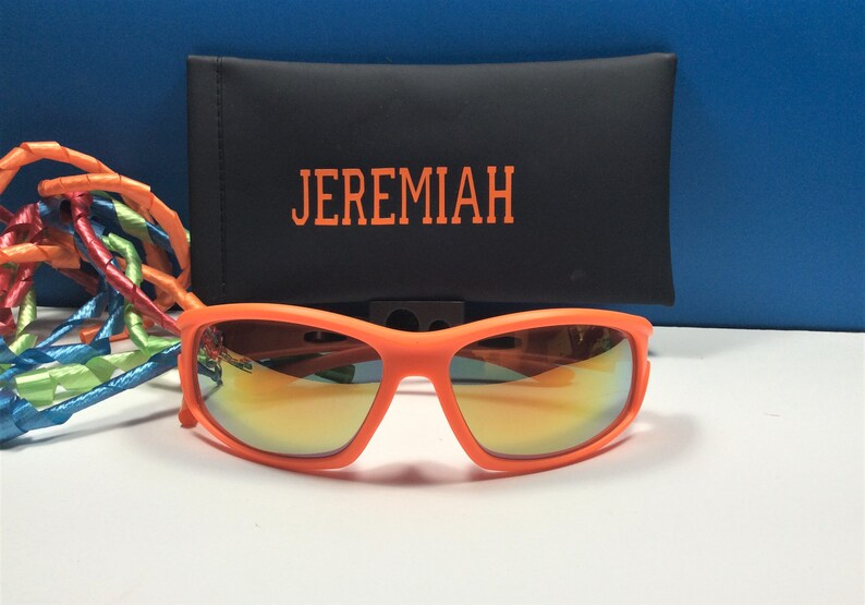 Puede incluir: Gafas de sol naranjas con lentes espejados y una funda negra con el nombre "JEREMIAH" bordado.