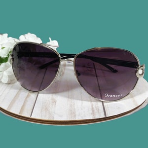 May include: Sunglasses with a silver frame and dark lenses, resting on a white wooden surface. The sunglasses have a curved design and the word "Frances" is printed on the lens. White flowers are in the background.