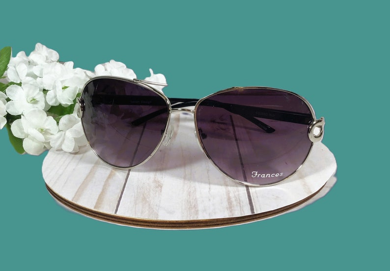 May include: Sunglasses with a silver frame and dark lenses, resting on a white wooden surface. The sunglasses have a curved design and the word "Frances" is printed on the lens. White flowers are in the background.