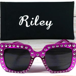 May include: Purple square sunglasses with rhinestones around the frame. The sunglasses are sitting on a black fabric case with the name "Riley" printed on it.
