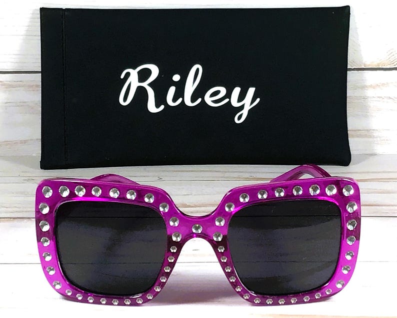May include: Purple square sunglasses with rhinestones around the frame. The sunglasses are sitting on a black fabric case with the name "Riley" printed on it.