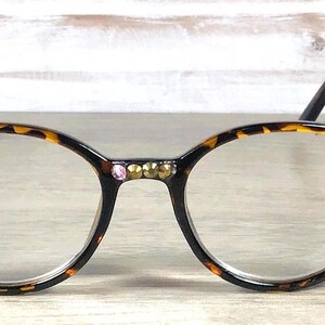 May include: Round eyeglasses with a tortoiseshell pattern frame. The frames are embellished with gold and pink rhinestones. The temples are a light brown color.