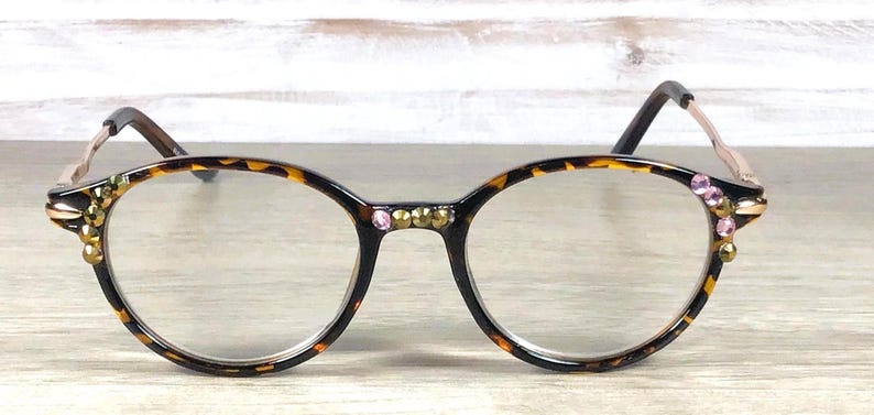May include: Round eyeglasses with a tortoiseshell pattern frame. The frames are embellished with gold and pink rhinestones. The temples are a light brown color.