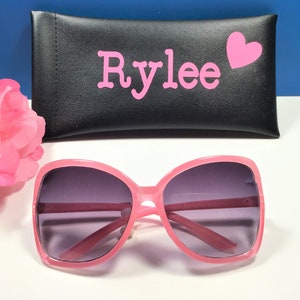 May include: Pink sunglasses with a matching case. The sunglasses have a large, square frame and dark lenses. The black case is personalized with the name "Rylee" in pink letters and a pink heart.