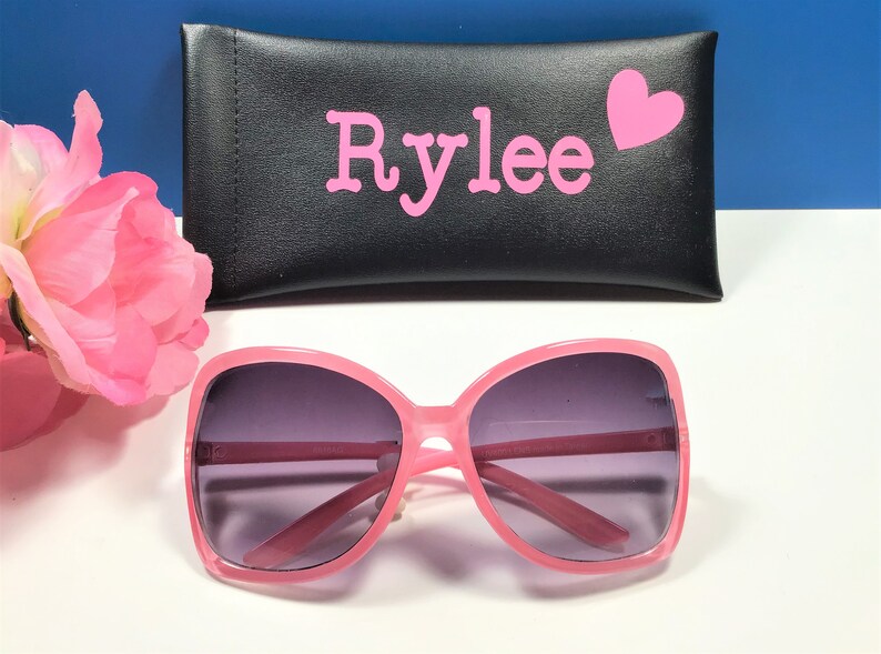 May include: Pink sunglasses with a matching case. The sunglasses have a large, square frame and dark lenses. The black case is personalized with the name "Rylee" in pink letters and a pink heart.