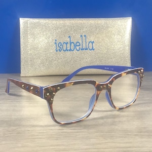 Puede incluir: Gafas de lectura con estampado de carey, detalles azules y tachuelas. Detr&aacute;s de las gafas hay un estuche dorado con el nombre "isabella" bordado en azul.