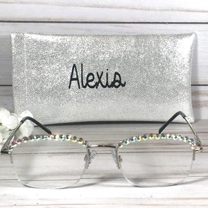 May include: Silver-toned semi-rimless glasses featuring iridescent rhinestones along the top of the frames. Behind the glasses sits a rectangular silver glitter case with the name Alexia printed in black script lettering on the front surface.