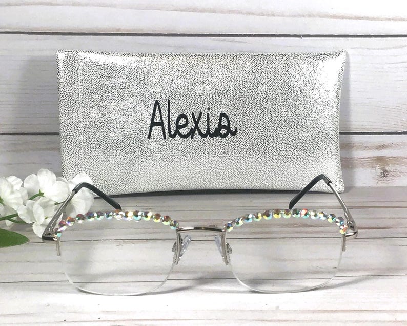 May include: Silver-toned semi-rimless glasses featuring iridescent rhinestones along the top of the frames. Behind the glasses sits a rectangular silver glitter case with the name Alexia printed in black script lettering on the front surface.