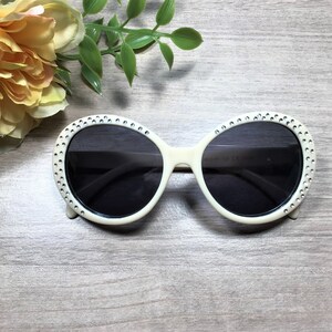 May include: White oval sunglasses with rhinestones around the frame and dark lenses.
