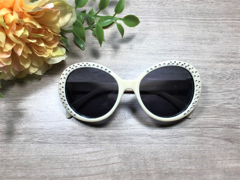 May include: White oval sunglasses with rhinestones around the frame and dark lenses.
