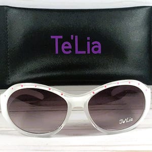 May include: White sunglasses with gradient lenses and small pink accents along the top frame. The right lens features the white text Te'Lia. Included is a black rectangular carrying case with the purple text Te'Lia printed on the front.