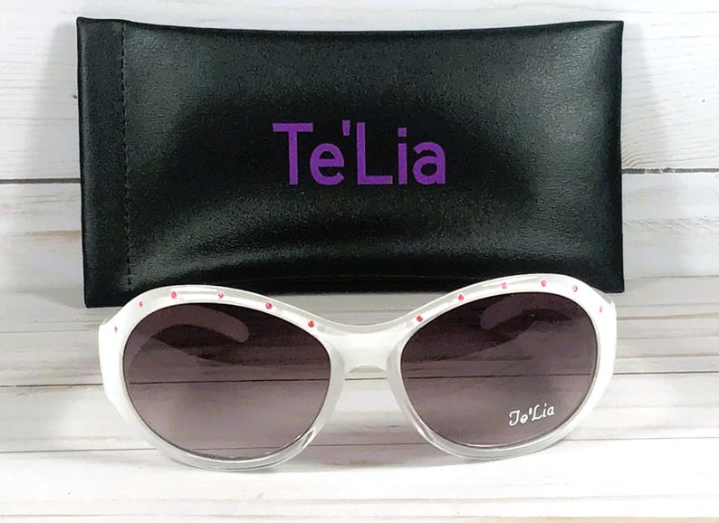 May include: White sunglasses with gradient lenses and small pink accents along the top frame. The right lens features the white text Te'Lia. Included is a black rectangular carrying case with the purple text Te'Lia printed on the front.