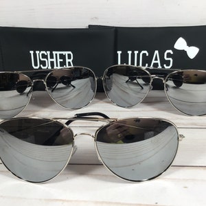 Może przedstawiać: Trzy pary srebrnych okular&oacute;w przeciwsłonecznych typu aviator z lustrzanymi szkłami. Okulary przeciwsłoneczne znajdują się przed dwoma czarnymi etui z nadrukowanymi nazwami "Usher" i "Lucas".