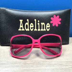 May include: Pink sunglasses with a black case that says "Adeline" with a pink flower.