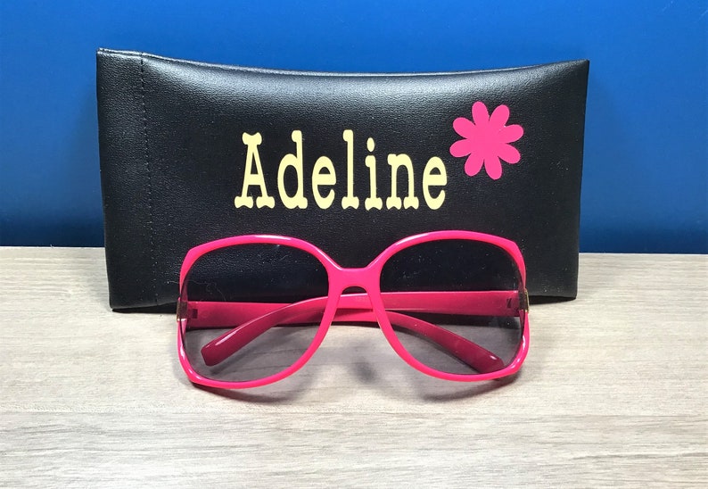 May include: Pink sunglasses with a black case that says "Adeline" with a pink flower.