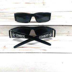 May include: Two pairs of black sunglasses. One pair is positioned above the other. The bottom pair has the word "COWBOYS" printed in white with a star logo on the temples. The sunglasses are on a white wooden surface.