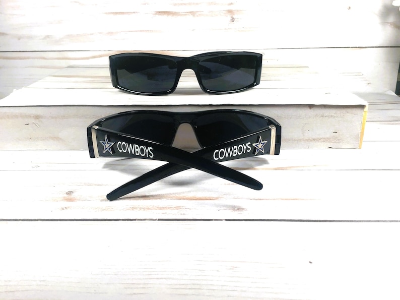 May include: Two pairs of black sunglasses. One pair is positioned above the other. The bottom pair has the word "COWBOYS" printed in white with a star logo on the temples. The sunglasses are on a white wooden surface.