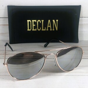 May include: Aviator sunglasses with a thin rose gold metal frame and reflective silver lenses. The sunglasses are positioned in front of a black rectangular case featuring the word DECLAN printed in gold lettering on the front surface.