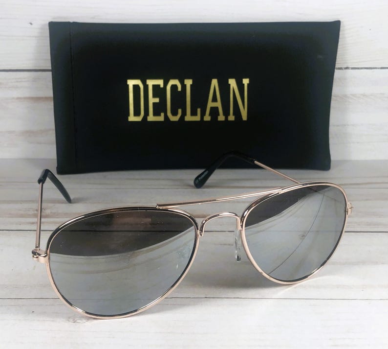 May include: Aviator sunglasses with a thin rose gold metal frame and reflective silver lenses. The sunglasses are positioned in front of a black rectangular case featuring the word DECLAN printed in gold lettering on the front surface.