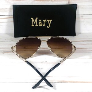Peut inclure: Lunettes de soleil aviateur dor&eacute;es avec des verres bruns et un d&eacute;tail de cha&icirc;ne dor&eacute;e tress&eacute;e sur les branches. Les lunettes de soleil sont dans un &eacute;tui noir avec le nom "Mary" brod&eacute; dessus.