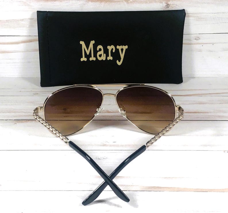 Peut inclure: Lunettes de soleil aviateur dor&eacute;es avec des verres bruns et un d&eacute;tail de cha&icirc;ne dor&eacute;e tress&eacute;e sur les branches. Les lunettes de soleil sont dans un &eacute;tui noir avec le nom "Mary" brod&eacute; dessus.