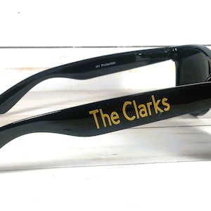 Puede incluir: Gafas de sol negras con el texto The Clarks en letras doradas en la patilla. La montura tiene un acabado brillante y lentes oscuras. El interior de la patilla incluye el texto UV Protection impreso en letras blancas pequeñas.