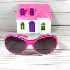 May include: Bright pink sunglasses with dark tinted lenses and small embedded rhinestones along the upper frame, positioned in front of a miniature white and pink toy house with a purple door and green window frames.