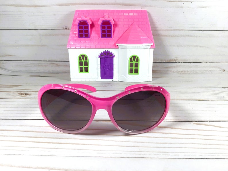 May include: Bright pink sunglasses with dark tinted lenses and small embedded rhinestones along the upper frame, positioned in front of a miniature white and pink toy house with a purple door and green window frames.