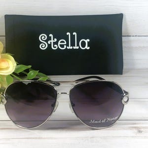 May include: Silver aviator sunglasses with gradient lenses, resting on a white wooden surface. The text "Maid of Honor" is printed on the lens. A black case with the name "Stella" in white script is behind the sunglasses.