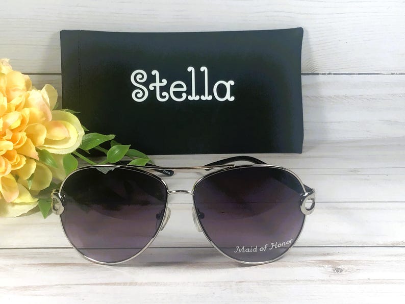 May include: Silver aviator sunglasses with gradient lenses, resting on a white wooden surface. The text "Maid of Honor" is printed on the lens. A black case with the name "Stella" in white script is behind the sunglasses.