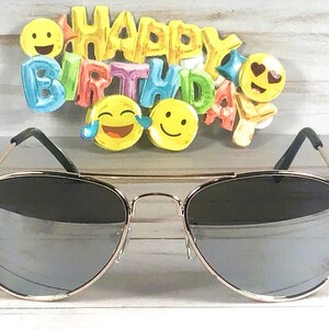 May include: Aviator sunglasses with thin gold-tone metal frames and reflective silver lenses, positioned in front of a colorful "HAPPY BIRTHDAY" sign featuring various yellow emoji faces including a laughing face and a heart-eyes face.