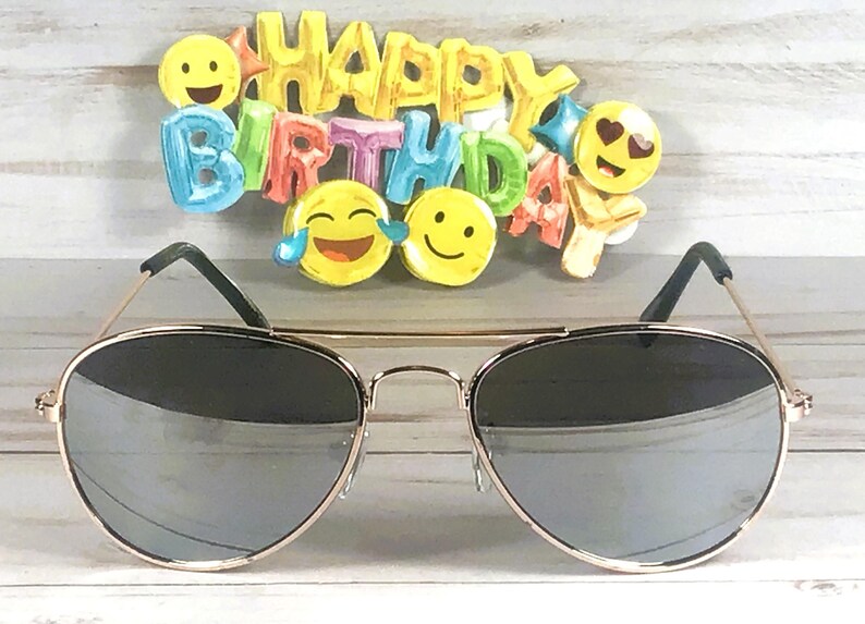 May include: Aviator sunglasses with thin gold-tone metal frames and reflective silver lenses, positioned in front of a colorful "HAPPY BIRTHDAY" sign featuring various yellow emoji faces including a laughing face and a heart-eyes face.