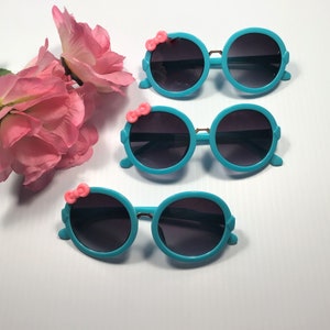 May include: Three pairs of turquoise blue sunglasses with pink bow accents. The sunglasses have round lenses and a plastic frame.