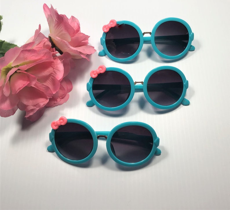 May include: Three pairs of turquoise blue sunglasses with pink bow accents. The sunglasses have round lenses and a plastic frame.
