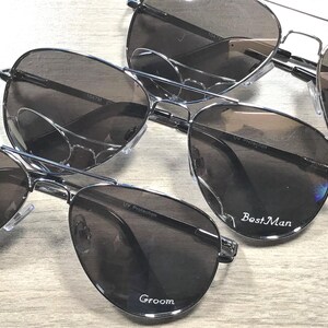 May include: Several pairs of aviator sunglasses with dark lenses and silver frames are arranged on a light wood surface. The sunglasses are labeled with the words "Groom", "Best Man", and "Groomsman".