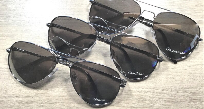 May include: Several pairs of aviator sunglasses with dark lenses and silver frames are arranged on a light wood surface. The sunglasses are labeled with the words "Groom", "Best Man", and "Groomsman".