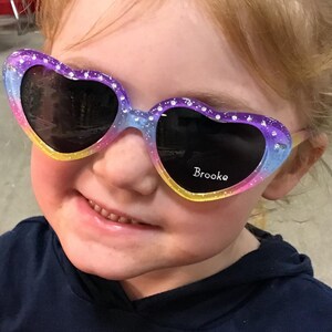 May include: A young girl wearing heart-shaped sunglasses with a rainbow glitter design. The sunglasses have the name "Brooke" written on the right lens.