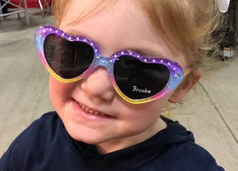May include: A young girl wearing heart-shaped sunglasses with a rainbow glitter design. The sunglasses have the name "Brooke" written on the right lens.