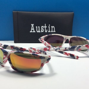 May include: A pair of white sunglasses with a red, white and blue camouflage pattern. The sunglasses have mirrored lenses and are folded in half. The sunglasses are sitting on a white surface next to a black case with the word "Austin" printed on it.
